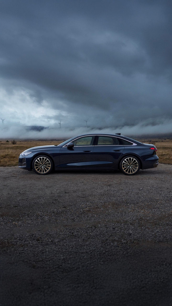 Elegante dunkelblaue Audi A6 Limousine auf einem Feldweg vor dramatischer Naturkulisse mit Nebel, Windrädern und dunklem Himmel. Das sportliche Fahrzeugdesign trifft auf eine eindrucksvolle Landschaft – Technik und Natur in perfekter Harmonie.