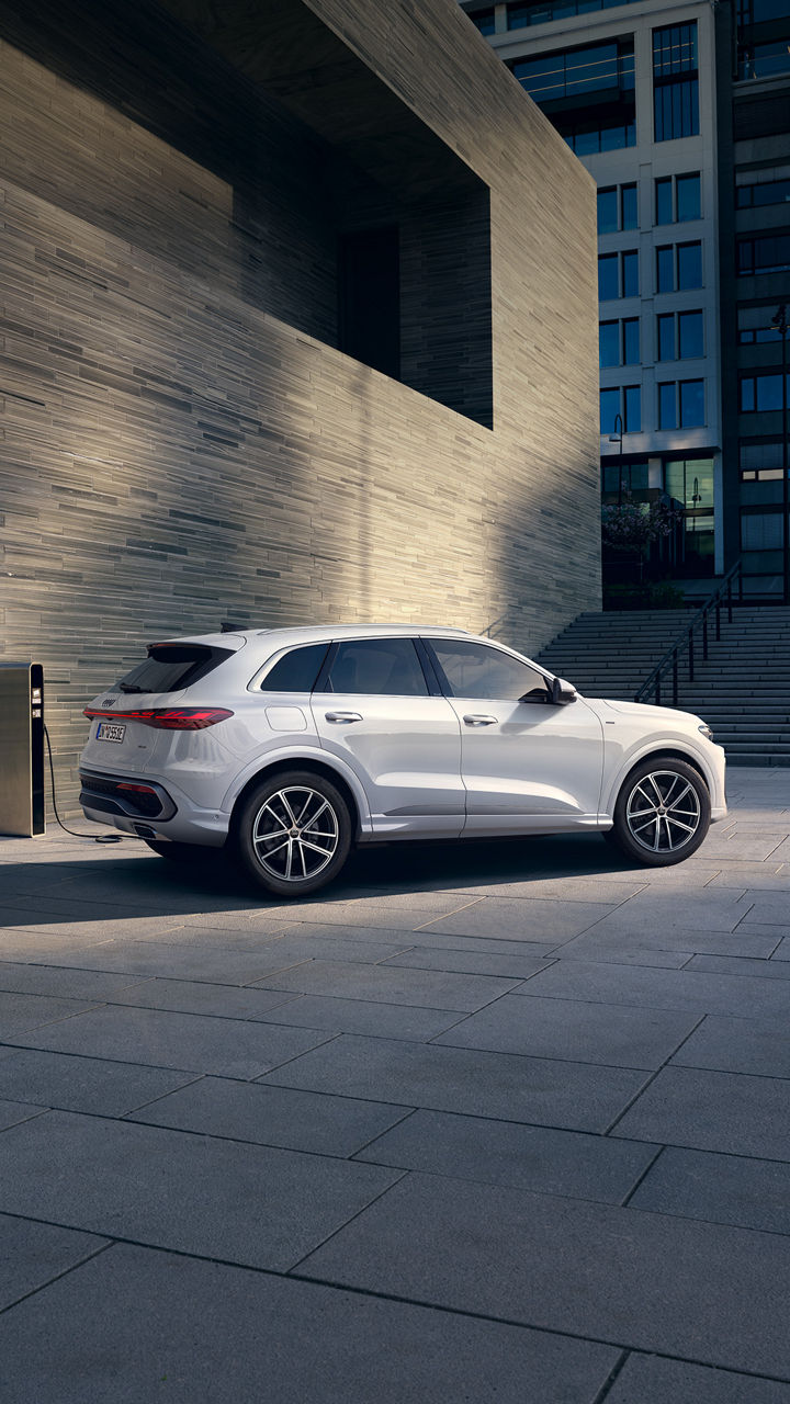 Weißer Audi Q5 e-hybrid auf einem Platz mit Ladesäule vor moderner Architektur.