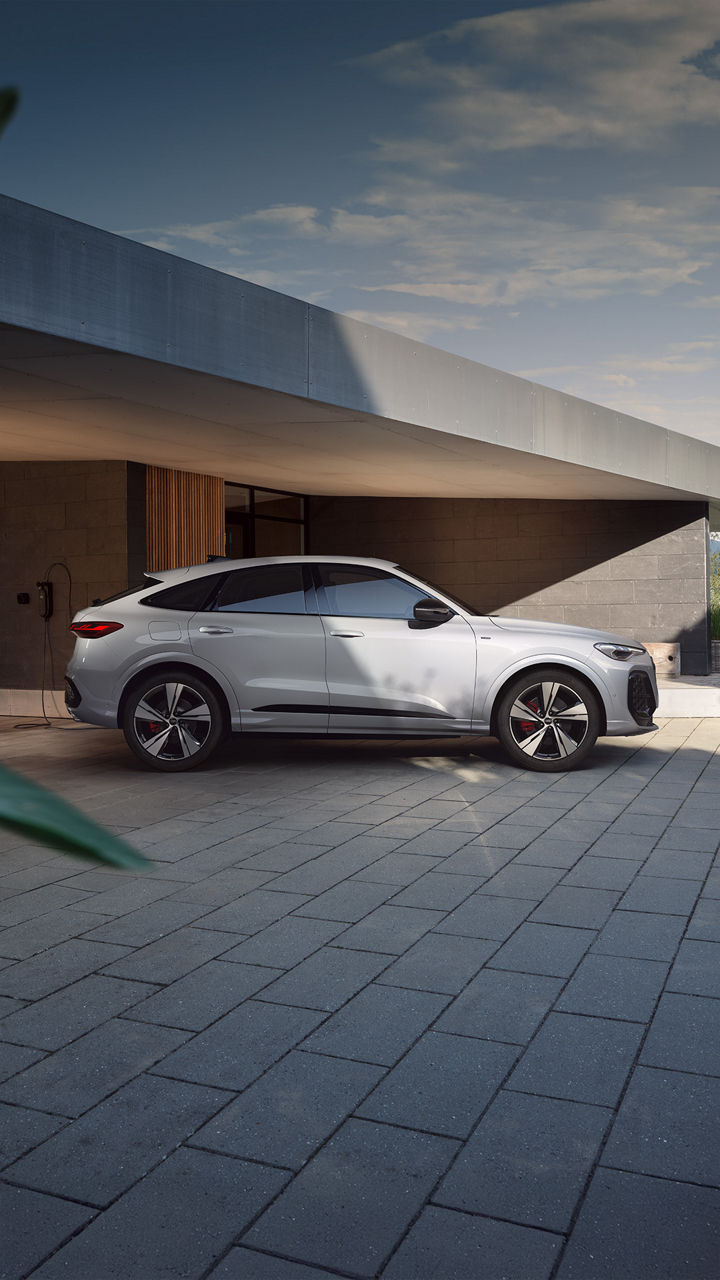 Weißer Audi Q5 Sportback e-hybrid vor moderner Architektur. Im Hintergrund ist eine Wall-Box an dem Wohnhaus zu sehen.