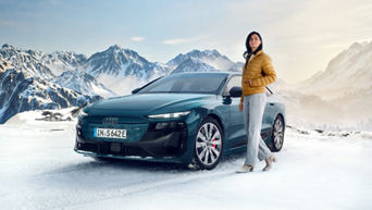 Eine Frau in einer gelben Winterjacke steht neben einem petrolfarbenen Audi S6 e-tron in einer Winterlandschaft. Im Hintergrund sind schneebedeckte Gipfel zu sehen.