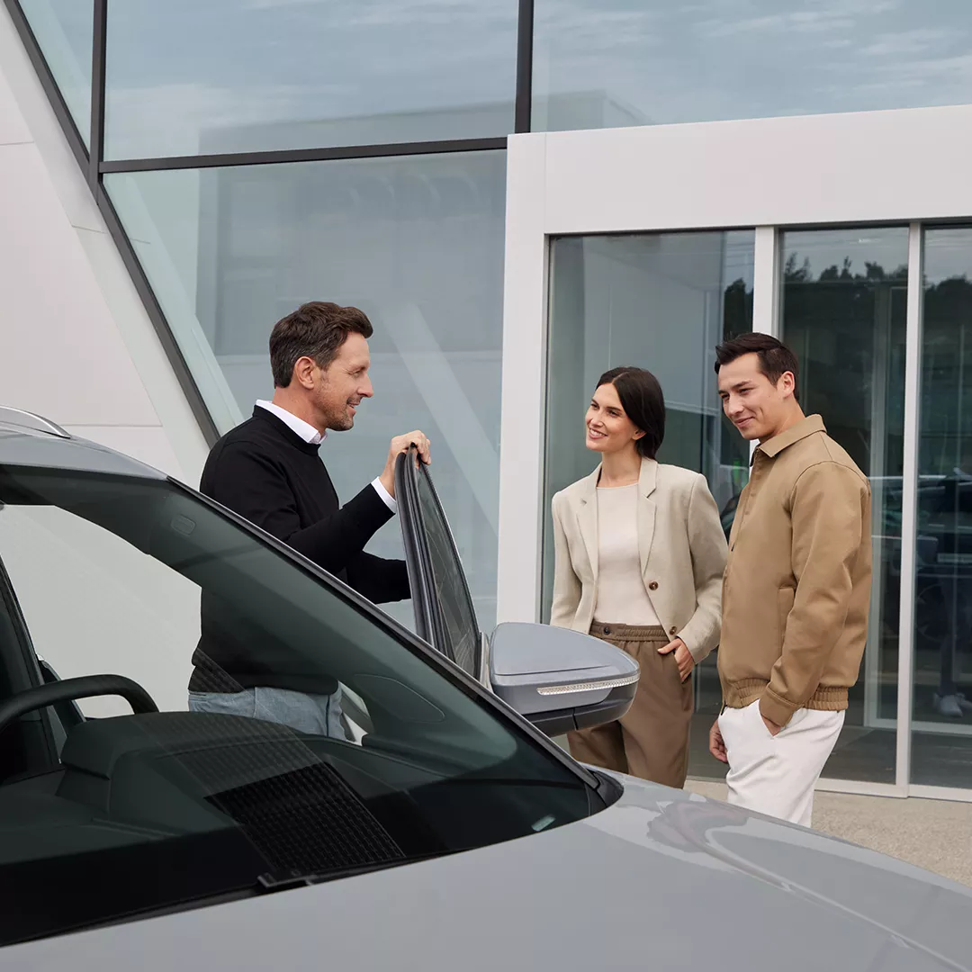 Eine Frau und zwei Männer stehen vor einem Autohaus von Audi. Ein Mann öffnet die Autotür eines Audi