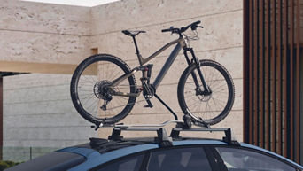 Mountainbike ist auf Dachträger eines blauen Audi montiert, im Hintergrund moderne Stein- und Holzarchitektur.