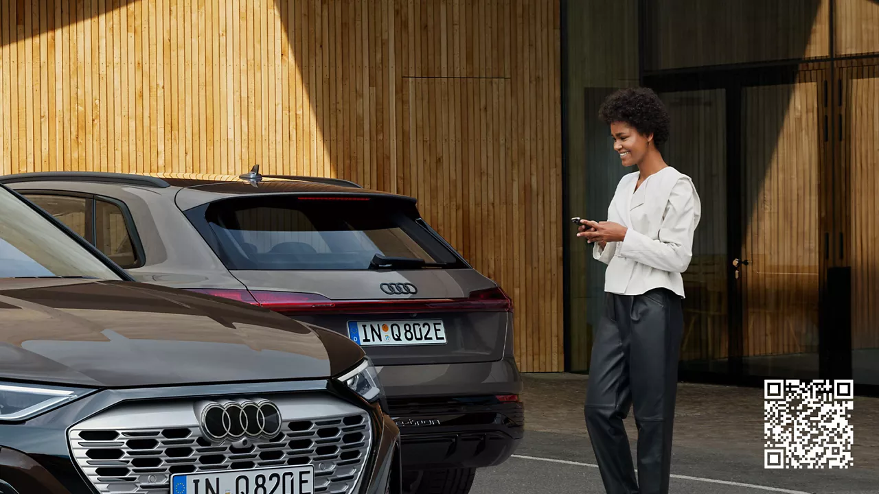 Deux voitures Audi sont garées côte à côte et une femme se tient à côté d'elles, les yeux rivés sur son téléphone.