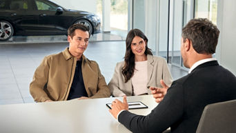 Un collaborateur Audi est en train de discuter avec un couple assis à son bureau. En arrière-plan, un véhicule Audi est exposé dans le showroom du concessionnaire.