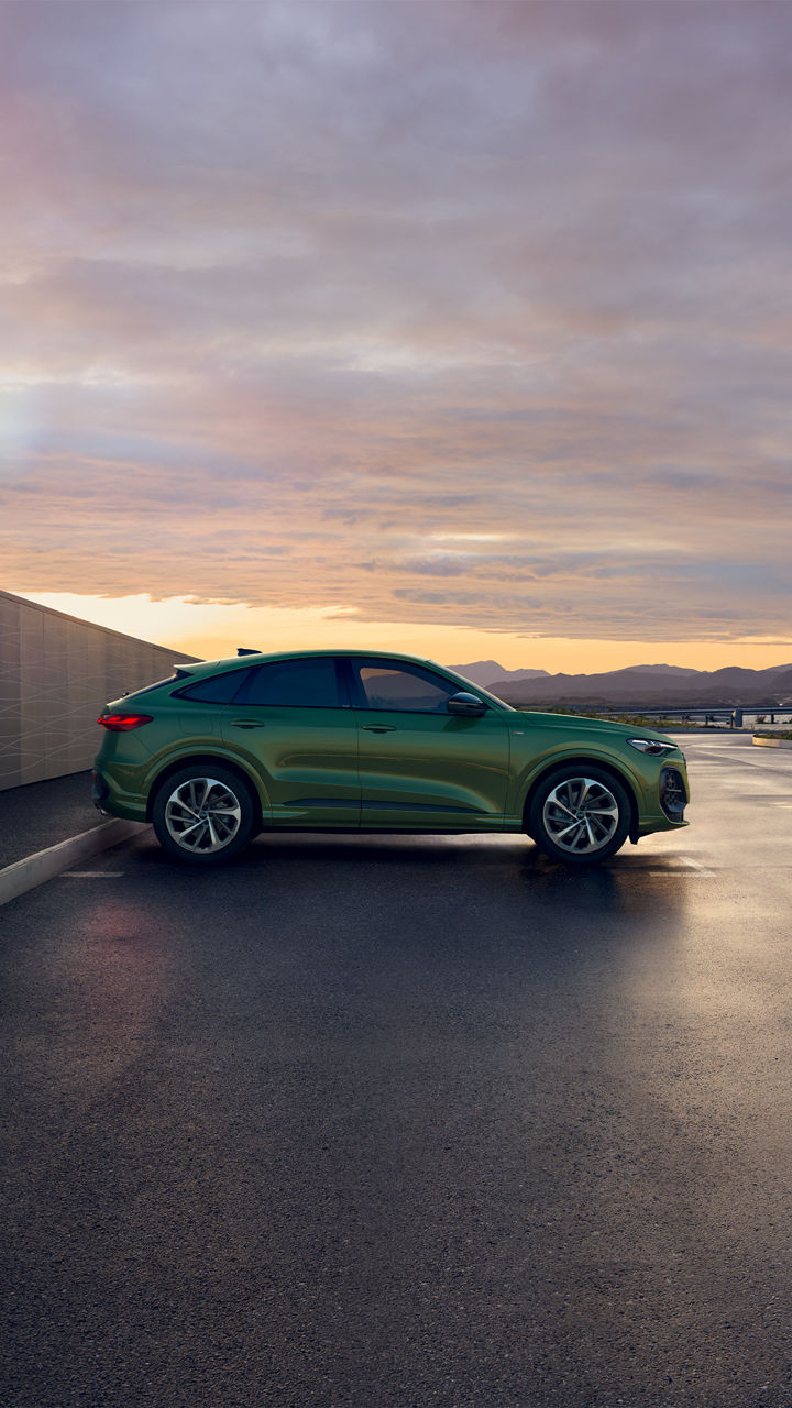 Seitenansicht eines grünen Audi Q5 Sportback, der bei Sonnenuntergang neben einer Mauer geparkt ist, mit einer bergigen Landschaft im Hintergrund.