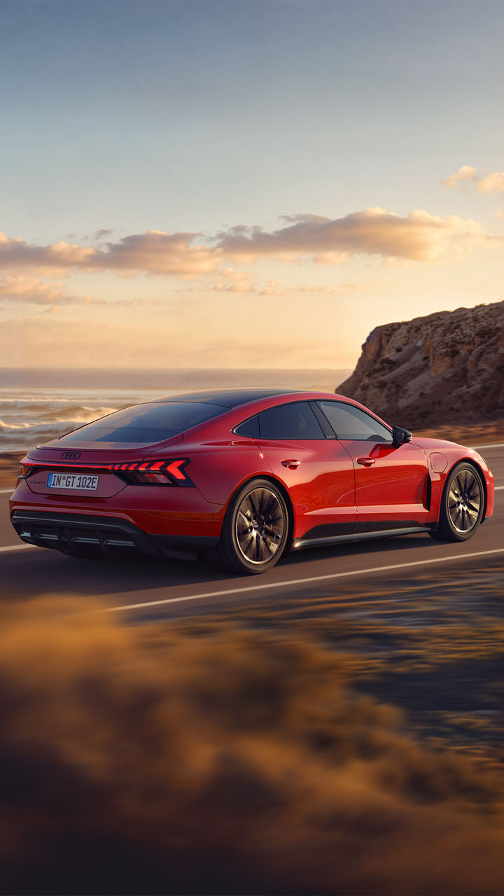 Heckansicht eines roten Audi e-tron GT quattro, der bei Sonnenuntergang auf einer Küstenstraße fährt, mit dem Meer und einer Klippe im Hintergrund.