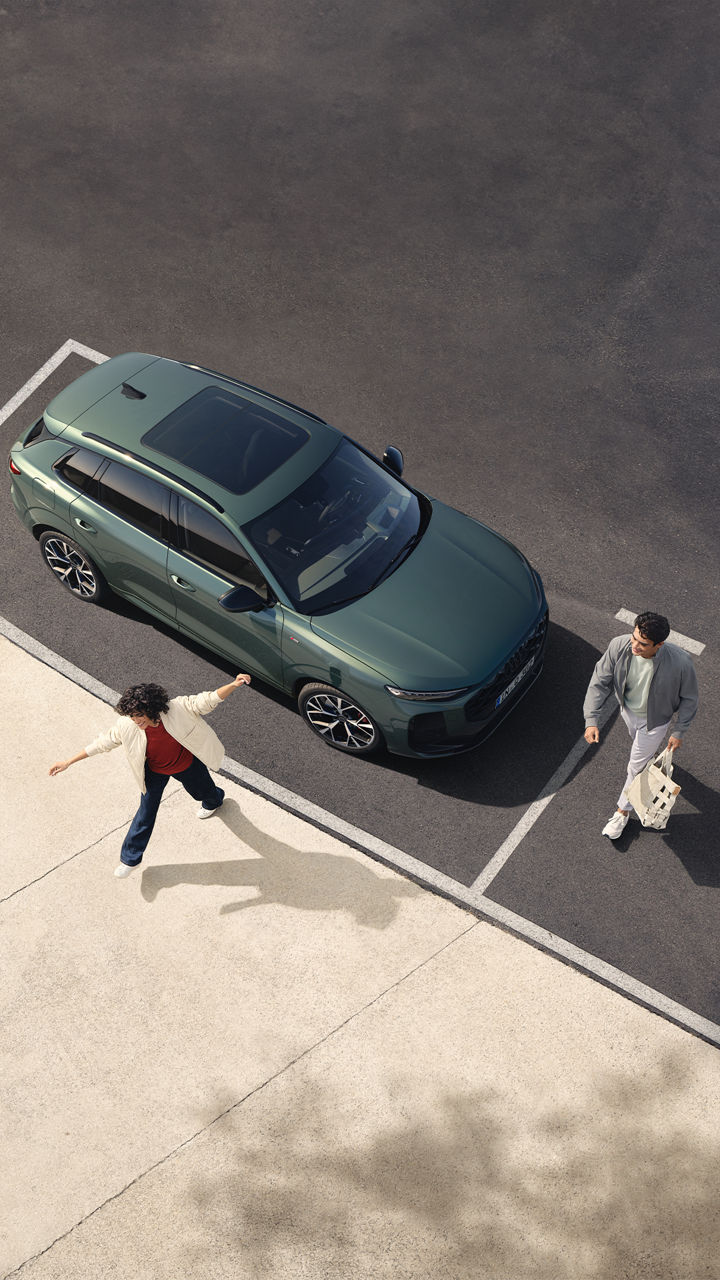 Von oben gesehen, stehen zwei Personen auf einem Parkplatz. Eine Frau, die Arme ausgestreckt, wendet sich einem Mann zu, der mit einer Tasche in der Hand geht. Ein dunkelgrüner Audi Q3 ist neben ihnen geparkt.