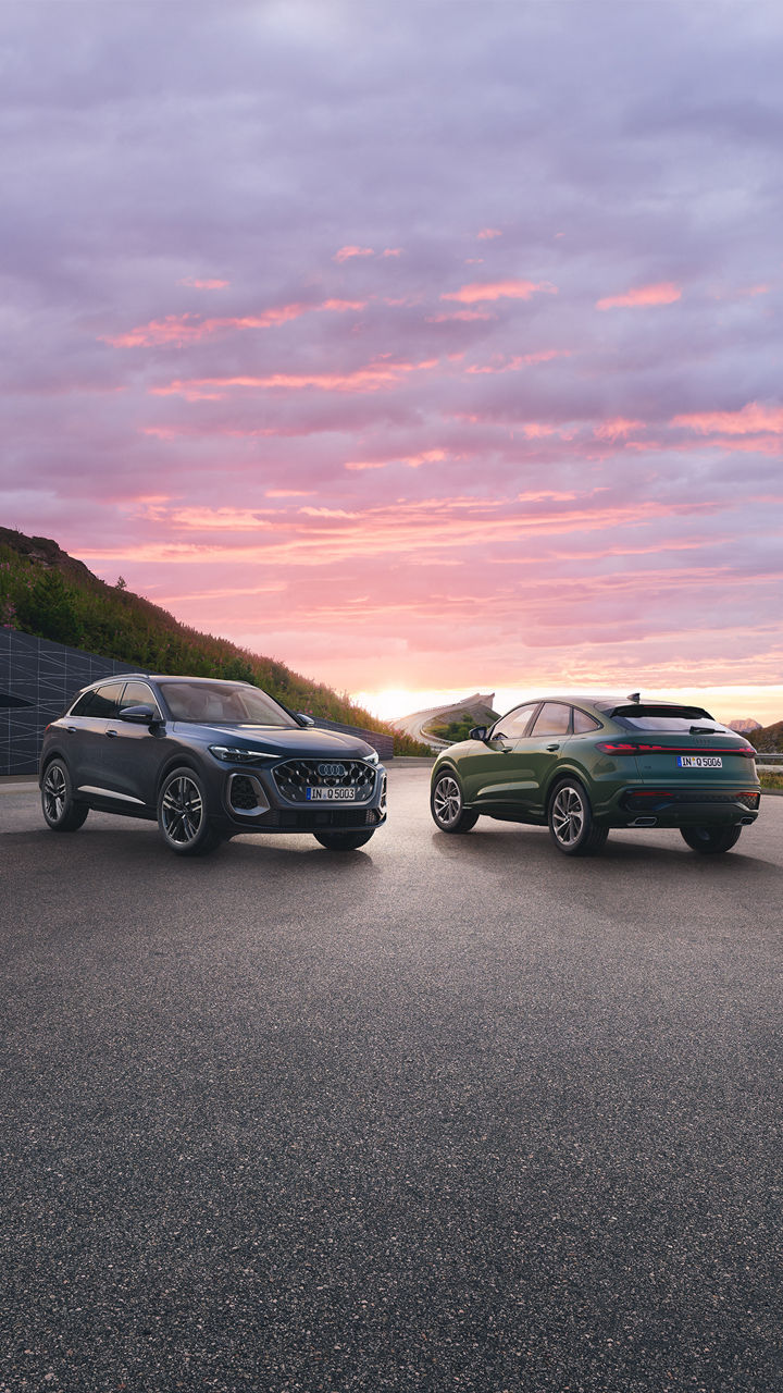 Zwei Audi Q5 stehen auf einer Bergstraße bei Sonnenuntergang. Der Himmel ist in sanften Farben gehalten und im Hintergrund sind grüne Hügel zu sehen.
