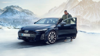 Berline Audi A6 noire ou bleu foncé sur un paysage de neige et de glace, avec un homme ouvrant la porte du conducteur et des montagnes enneigées en arrière-plan.