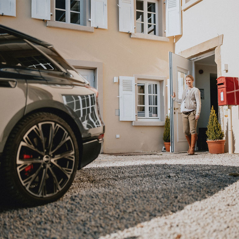 Theresa Baumgärtner verlässt durch eine Tür das Haus, an dem ein großer roter Briefkasten hängt. Im Vordergrund sieht man das Hinterrad des geparkten Audi A6 e-tron.