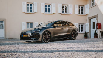 Un Audi A6 e-tron est garé sur un sol en gravier devant une maison beige aux volets blancs.