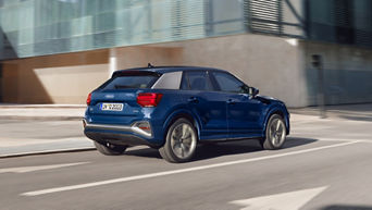 Un Audi Q2 bleu foncé roule sur une route devant un bâtiment moderne.
