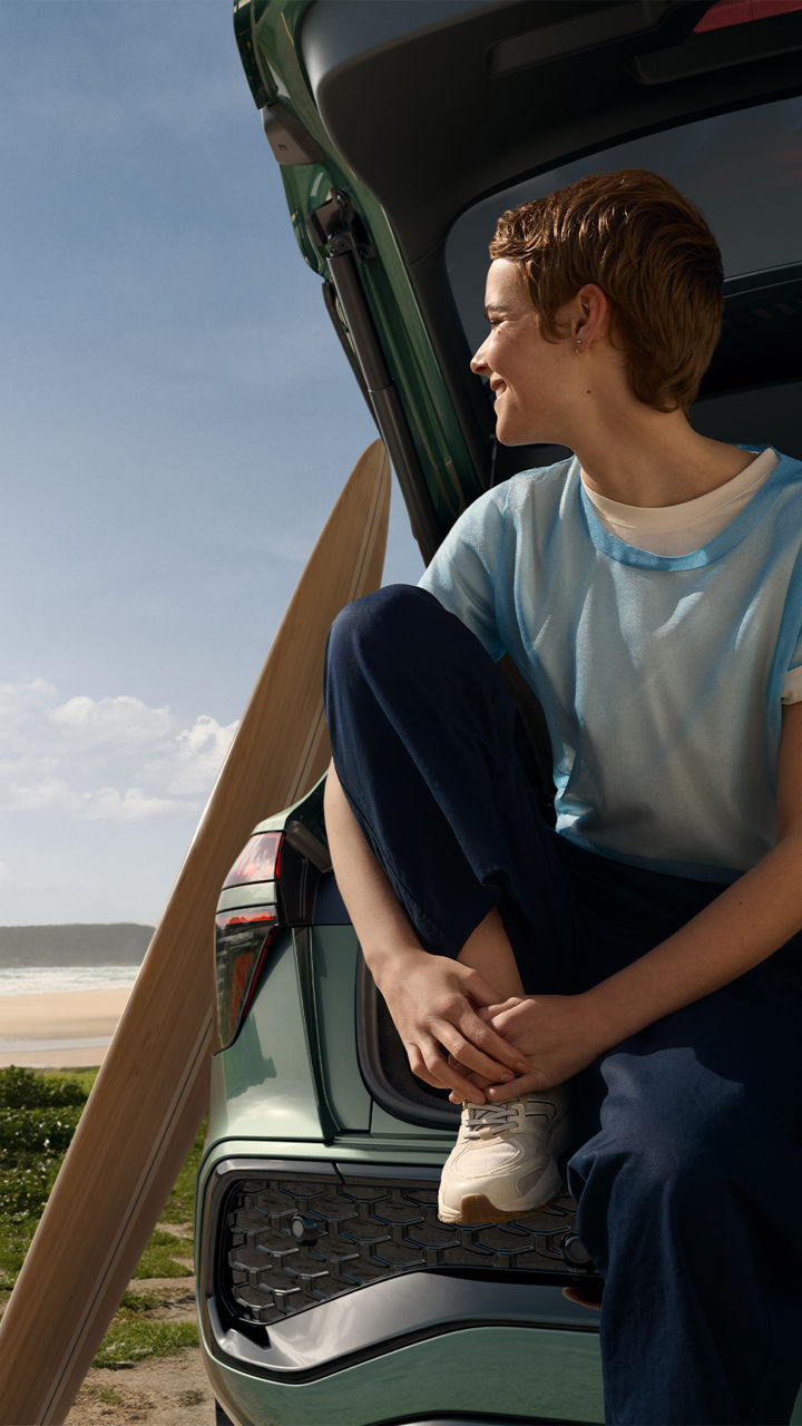 Une femme aux cheveux courts assise dans le coffre ouvert d'une voiture, regardant vers une plage ensoleillée avec l'océan en arrière-plan.