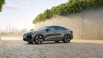 Un Audi Q6 e-tron Sportback gris foncé est garé sur une place pavée devant un mur en béton avec de la verdure et une ligne d'horizon en arrière-plan.