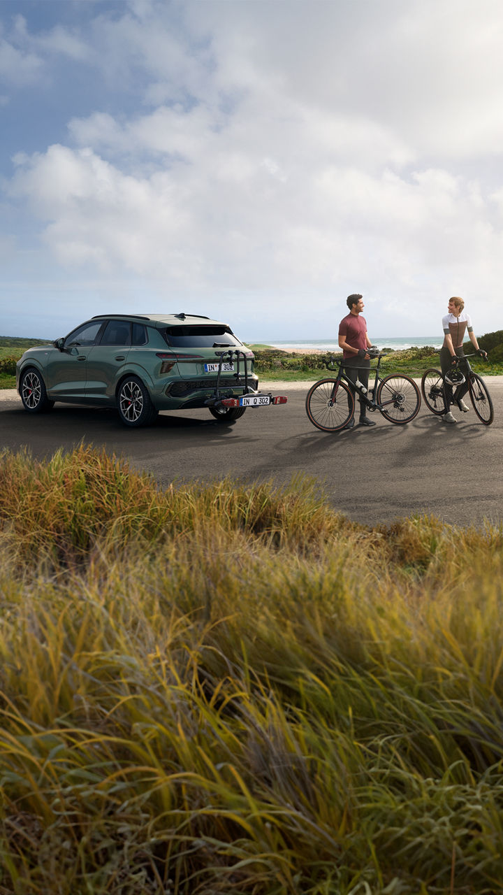 Ein grüner Audi Q4 E-Tron Sportback mit Fahrradträger steht auf einem Parkplatz am Meer, neben dem Auto stehen zwei Personen mit Fahrrädern.