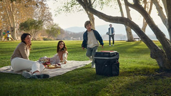 Eine Familie macht ein Picknick im Park: Eine Frau sitzt auf einer Decke, ein Mädchen liegt daneben, ein Junge läuft auf eine Kühlbox zu, und ein Mann ist im Hintergrund zu sehen.