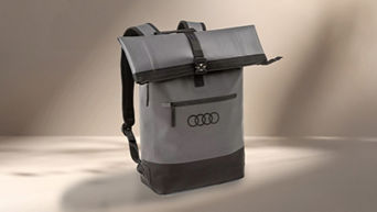 Ein grauer Roll-Top-Rucksack mit schwarzem Boden und einem Audi-Logo ist auf einem beigen Hintergrund mit Schatteneffekten dargestellt.