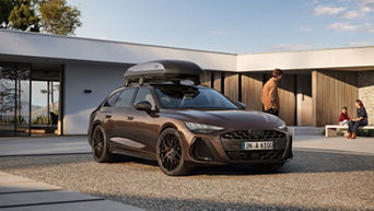  Ein brauner Audi A5 Sportback mit Dachbox steht auf einem Kiesweg vor einem modernen Haus, während zwei Personen im Hintergrund draußen sitzen und eine weitere Person ins Haus geht.