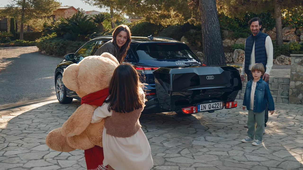 Eine Familie steht um den geöffneten Kofferraum eines dunklen Audi Q5, wobei ein Kind einen großen Teddybär hält.