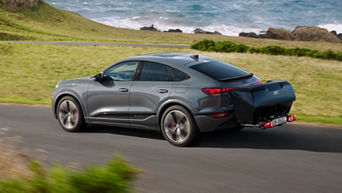 Grauer Audi Q6 Sportback fährt auf Straße mit einer Heckbox montiert. Im Hintergrund sieht man das Meer.