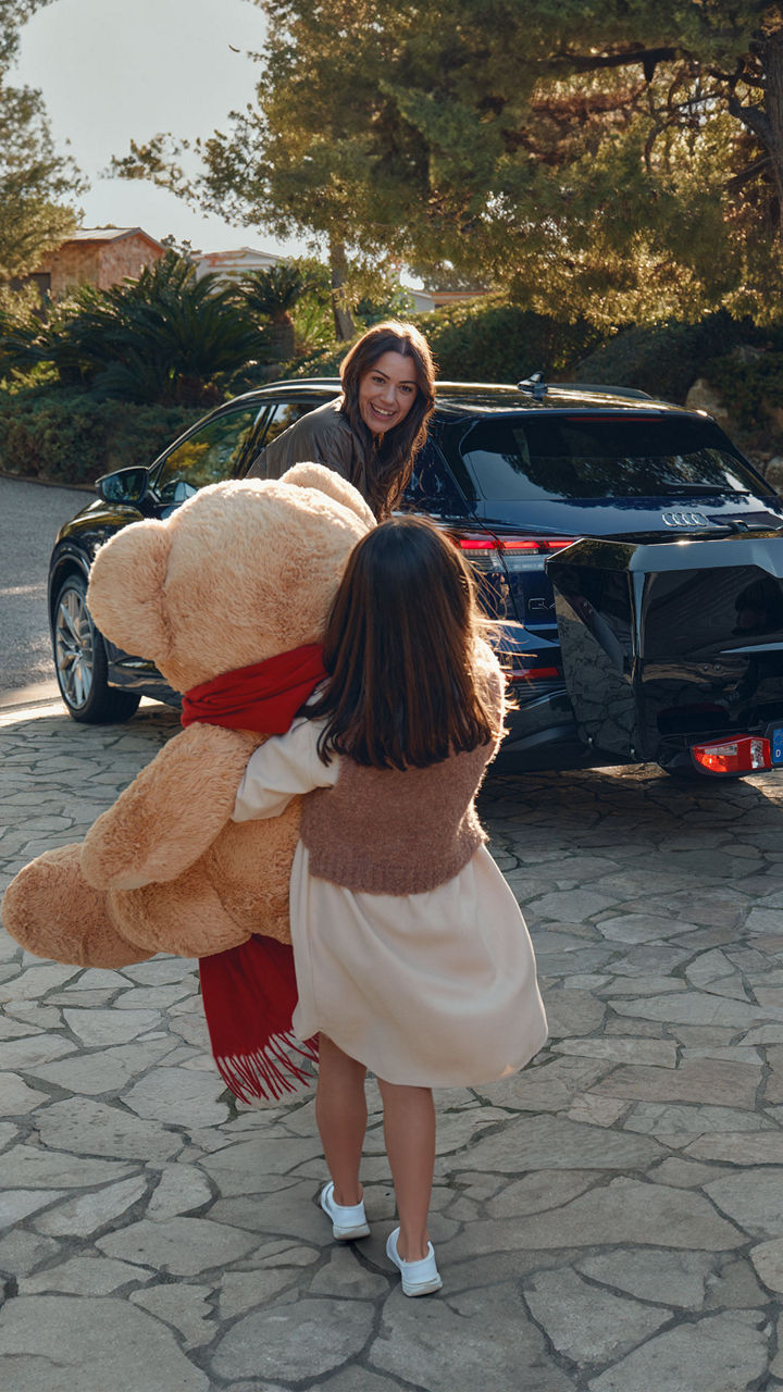Une famille se tient devant une Audi noire avec boîte arrière. Une petite fille s'approche de la famille et porte un très gros ours en peluche vers la voiture.