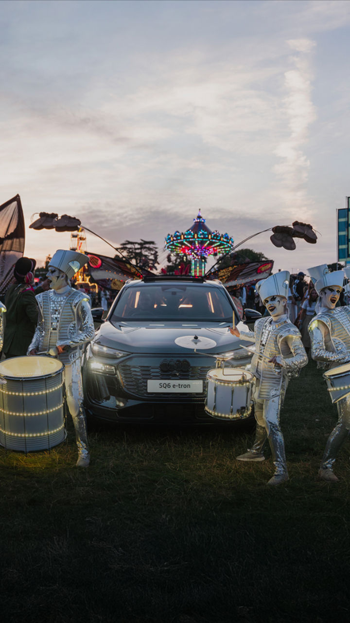 Performers in metallic costumes play illuminated drums beside an Audi SQ6 e-tron at an outdoor event. Large winged structures and colorful flags adorn the background.