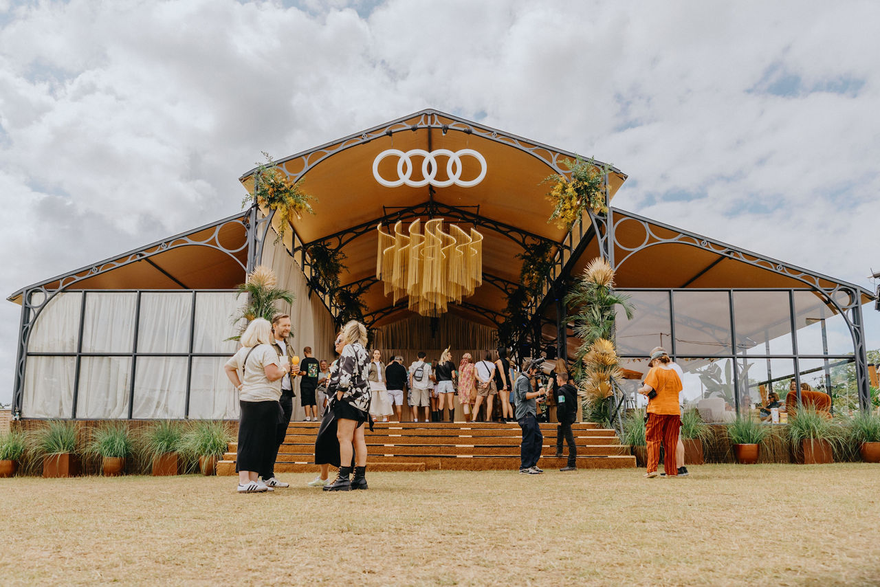 A modern event tent adorned with greenery, featuring an Audi logo, bustling with people on a grassy area under a cloudy sky.