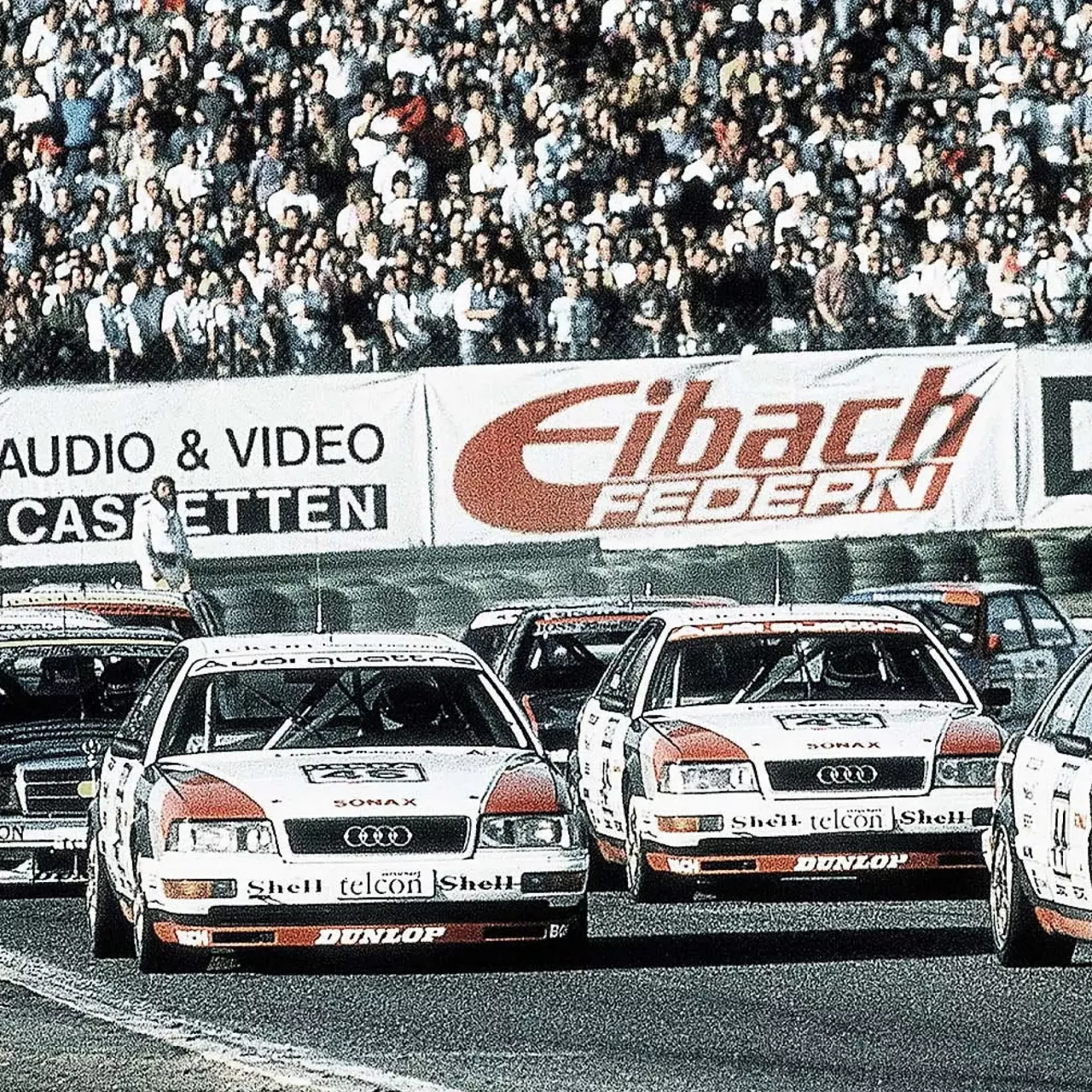 Two Audi racing cars among other racing cars on a track with a crowd in the background and advertising banners above