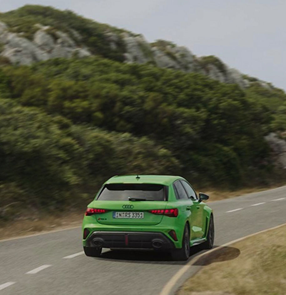 A green Audi driving around a scenic curve, surrounded by lush greenery and rocky hills.