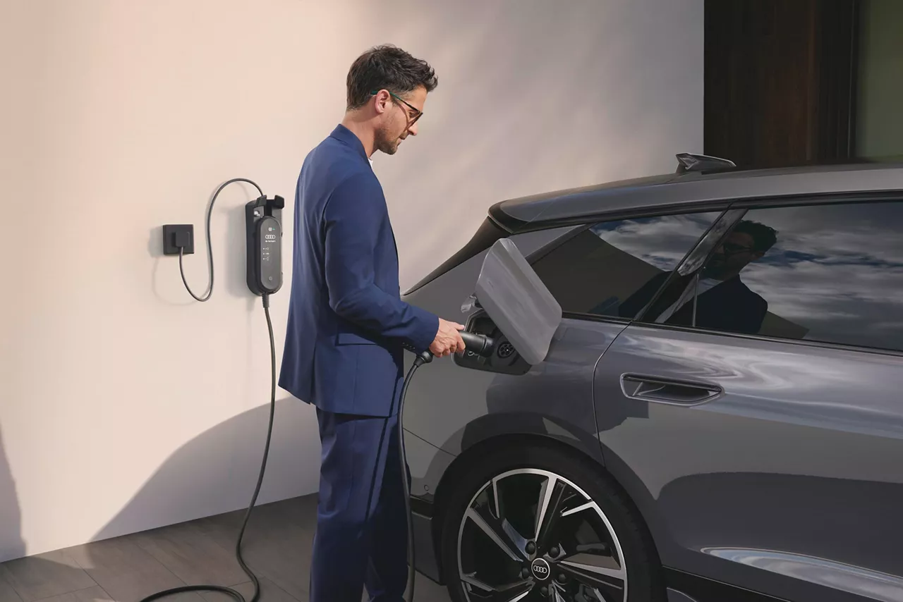 A man in a blue suit plugs an electric Audi into a charging station on a modern home exterior.