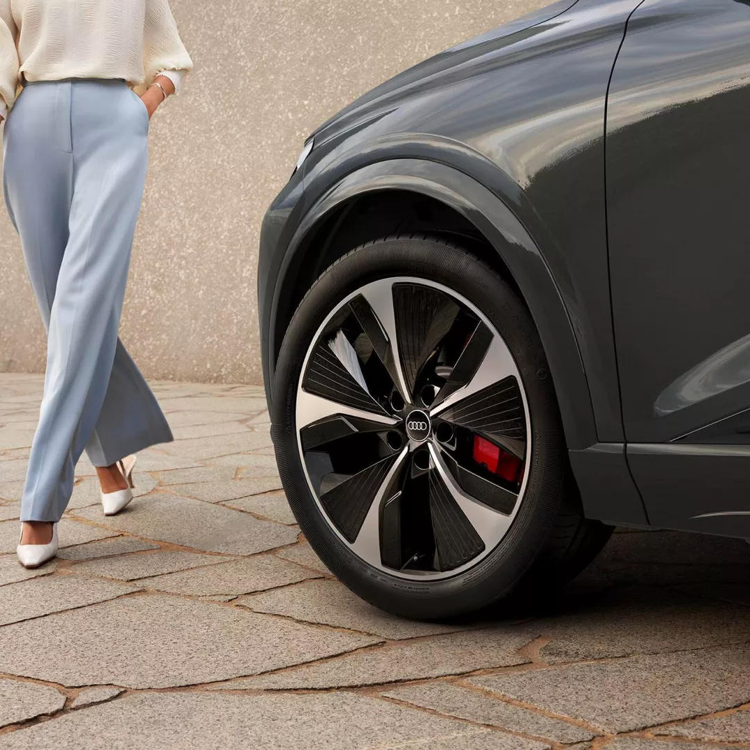 A parked Audi electric vehicle with focus on the front left of the vehicle and womans legs with long pants and white heels