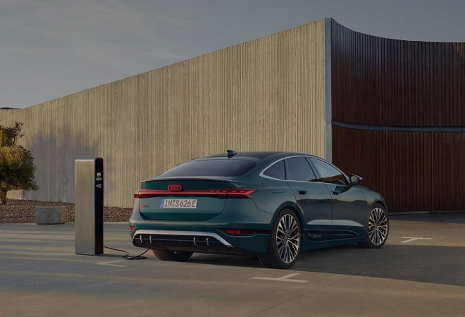 A man walking towards us with a parked black Audi electric vehicle on charge in the background 