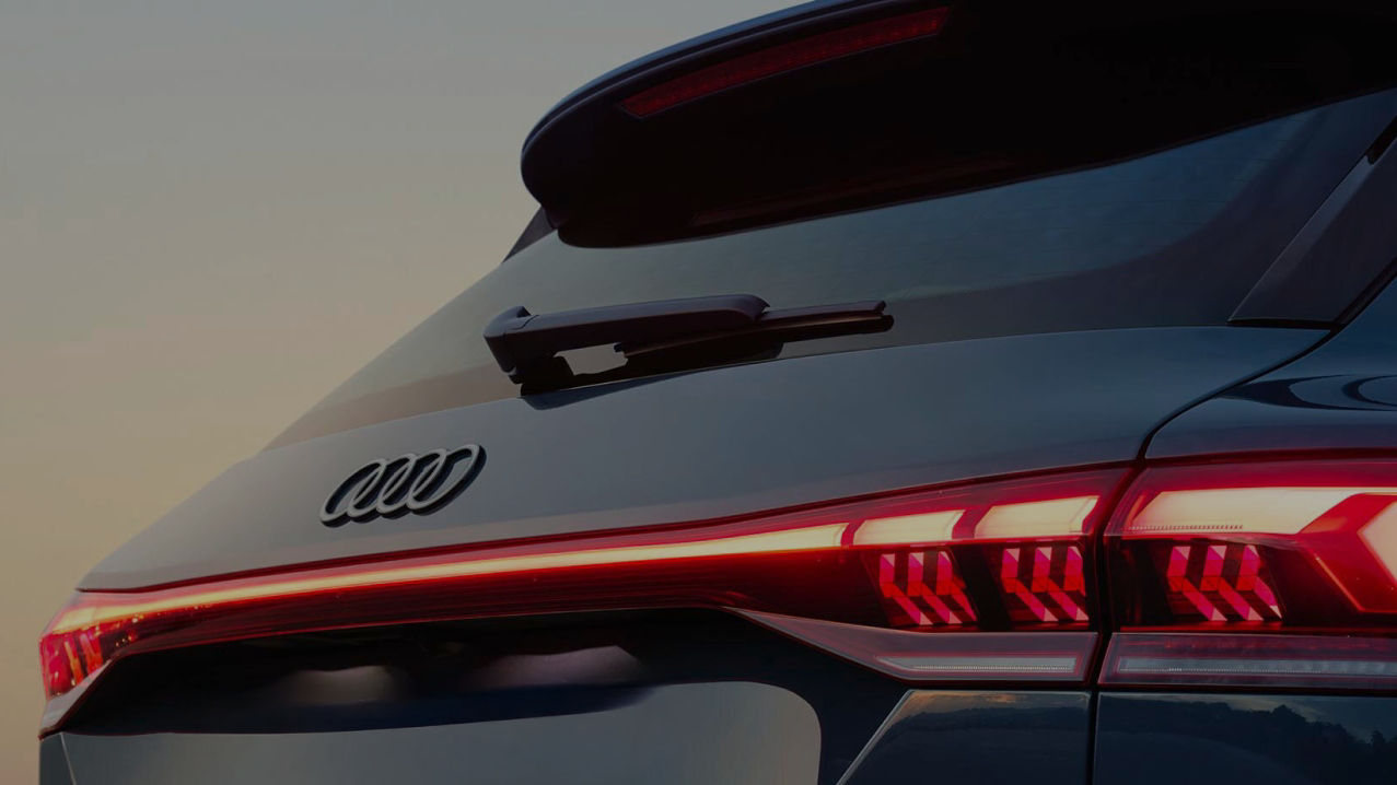 Close-up of the rear of an Audi car, showcasing its logo, sleek design, and illuminated taillights against a soft sunset backdrop.