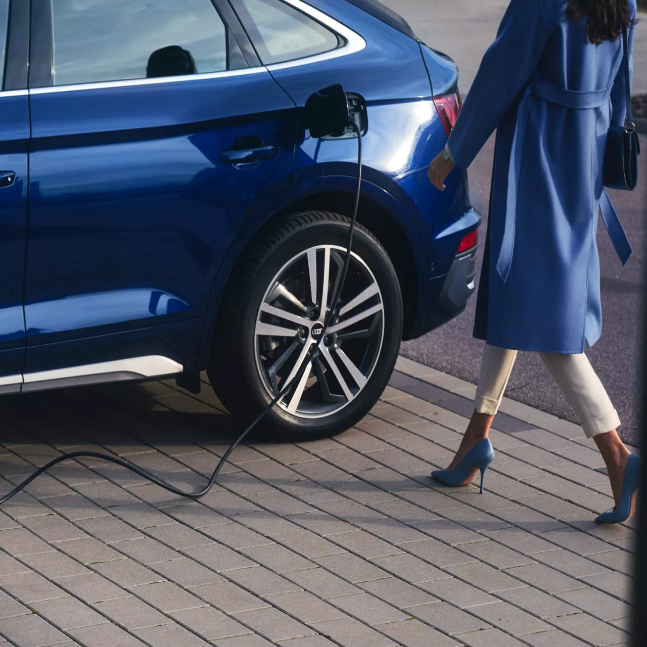 Woman in blue trench coat, approaching a dark blue Audi electric vehicle