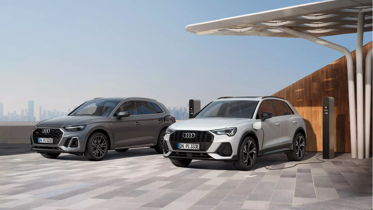 Two Audi vehicles parked side by side, one charging at a station, with a cityscape in the background