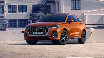 An orange Audi Q3 SUV parked in an urban setting, showcasing its sporty design against a backdrop of modern architecture.