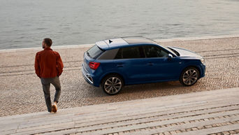 A person walks down steps towards a blue Audi Q2 parked by the water, surrounded by cobblestone pathways.