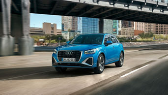 A blue Audi Q2 drives on a city street, with modern buildings and an overpass in the background, showcasing its sleek design.