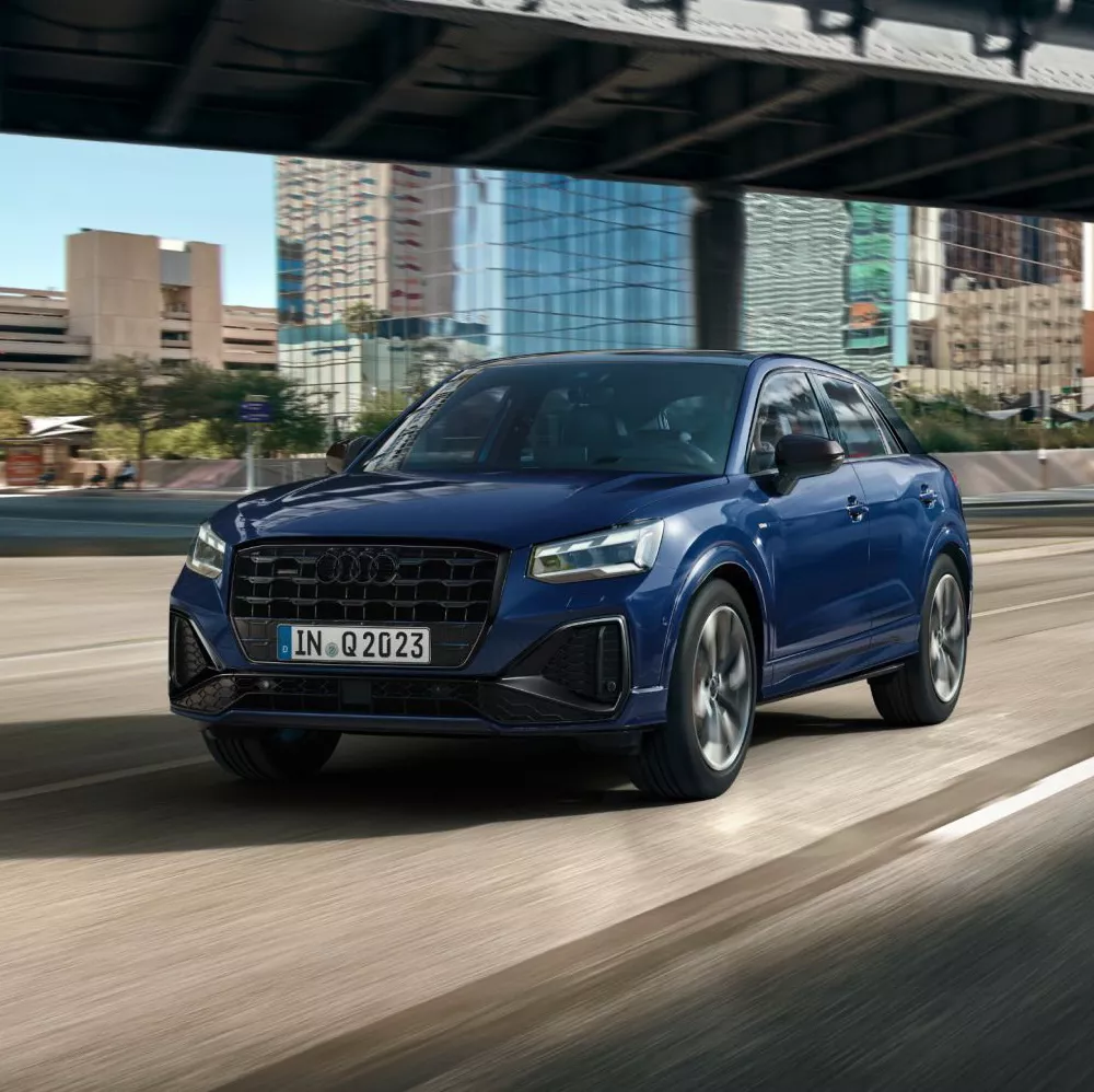 A blue Audi Q2 SUV driving on a sunlit road, with modern buildings reflecting in the background.