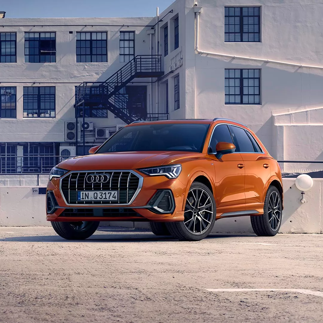 An orange Audi Q3 SUV parked in front of a modern, industrial-style building with large windows and a staircase.