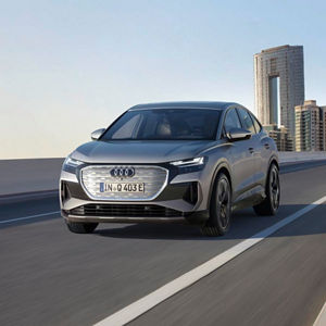 A silver Audi Q4 e-tron drives on a modern highway, with urban skyscrapers in the background under a clear sky.