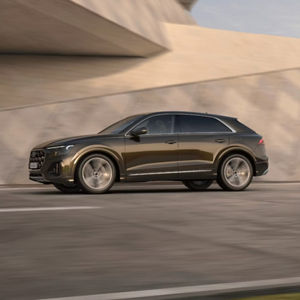 A dark brown Q8 SUV TFSI e speeds past a modern architectural backdrop, capturing motion and luxury in an urban setting.