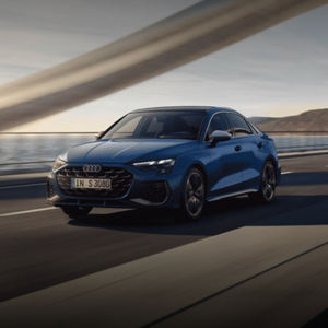 A blue Audi S3 speeds along a winding road with a scenic backdrop of water and hills.