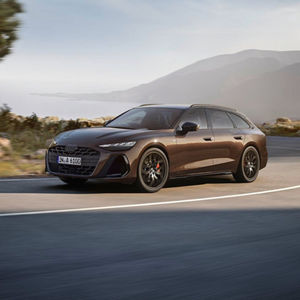 A brown Audi A6 Avant drives along a winding coastal road, showcasing its modern design against a scenic backdrop.