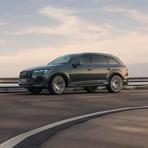 A grey Audi Q7 SUV driving on a curvy road at sunset, showcasing its modern design against a scenic backdrop.