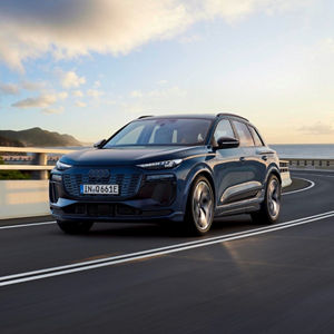 A blue Audi Q6 e-tron electric SUV driving on a coastal road with mountains and a sunset in the background.
