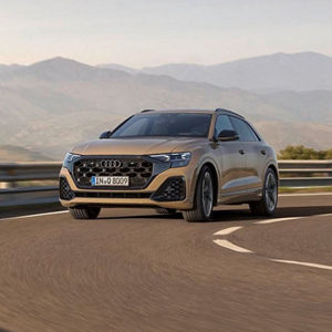 An Audi Q8 SUV in a sleek bronze color driving on a winding road, with mountains in the background under a clear sky.