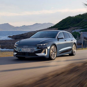 A sleek Audi A6 e-tron, in silver and black, drives along a coastal road with mountains in the background under a clear blue sky.