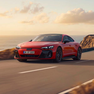 A red Audi e-tron GT drives along a coastal road, with a scenic ocean backdrop and gentle clouds in the sky.