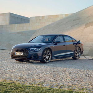 A black Audi A8 TFSI e parked on a cobblestone surface, charging alongside a modern architectural background at sunset.
